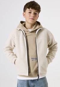 Chaqueta de forro polar color crema con cremallera frontal, dos bolsillos laterales y una textura suave, superpuesta a una sudadera beige con detalles de texto.