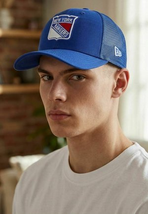 Junger Mann, der eine blaue New York Rangers Kappe und ein weißes Shirt mit Rundhalsausschnitt trägt, leicht nach links blickt und einen neutralen Gesichtsausdruck zeigt, drinnen.