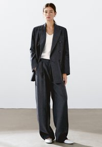 Completo sartoriale a righe scure con blazer doppiopetto e pantaloni a gamba larga, abbinato a un top bianco aderente e a scarpe bianche semplici.