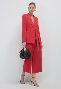 Blazer rosso sartoriale e pantaloni a gamba larga, cinched in vita con una cintura. Borsa nera e tacchi a strappo neri completano l'outfit.