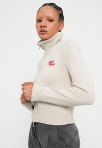 Pull en maille beige à col roulé avec fermeture éclair, poignets et ourlet côtelés. Présente un texte brodé rouge sur le côté gauche.