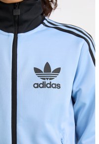 Šviesiai mėlynas Adidas treniruoklių džemperis su juodu užtrauktuku, juodu apykakle, juodomis juostelėmis ant pečių ir Adidas trefoilio logotipu ant krūtinės.