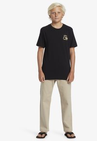 Zwarte katoenen t-shirt met een gouden logo aan de voorkant, gecombineerd met beige rechte broek en zwarte slippers. Casual fit, minimalistisch design.