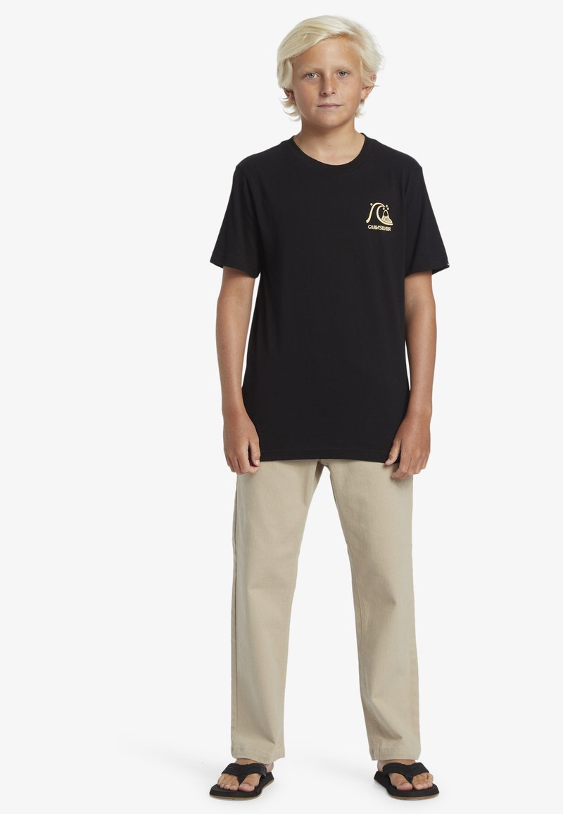 Zwarte katoenen t-shirt met een gouden logo aan de voorkant, gecombineerd met beige rechte broek en zwarte slippers. Casual fit, minimalistisch design.