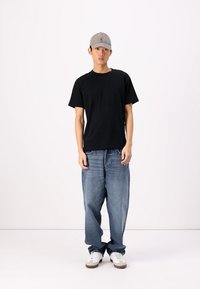 Svart bomulls-t-skjorte, løse blå denimjeans, grå caps og hvite sneakers med brune detaljer, stående mot en ensfarget bakgrunn.