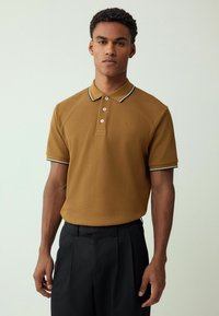 Camisa polo marrón con tejido texturizado, tapeta de tres botones y acentos de color blanco y marino en el cuello, combinada con pantalones negros.