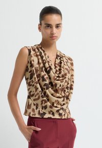 Vrouw in een mouwloze beige top met bruine luipaardprint en gedrapeerde halslijn, gecombineerd met hooggewaisted bordeauxrode broek, staand tegen een effen achtergrond.