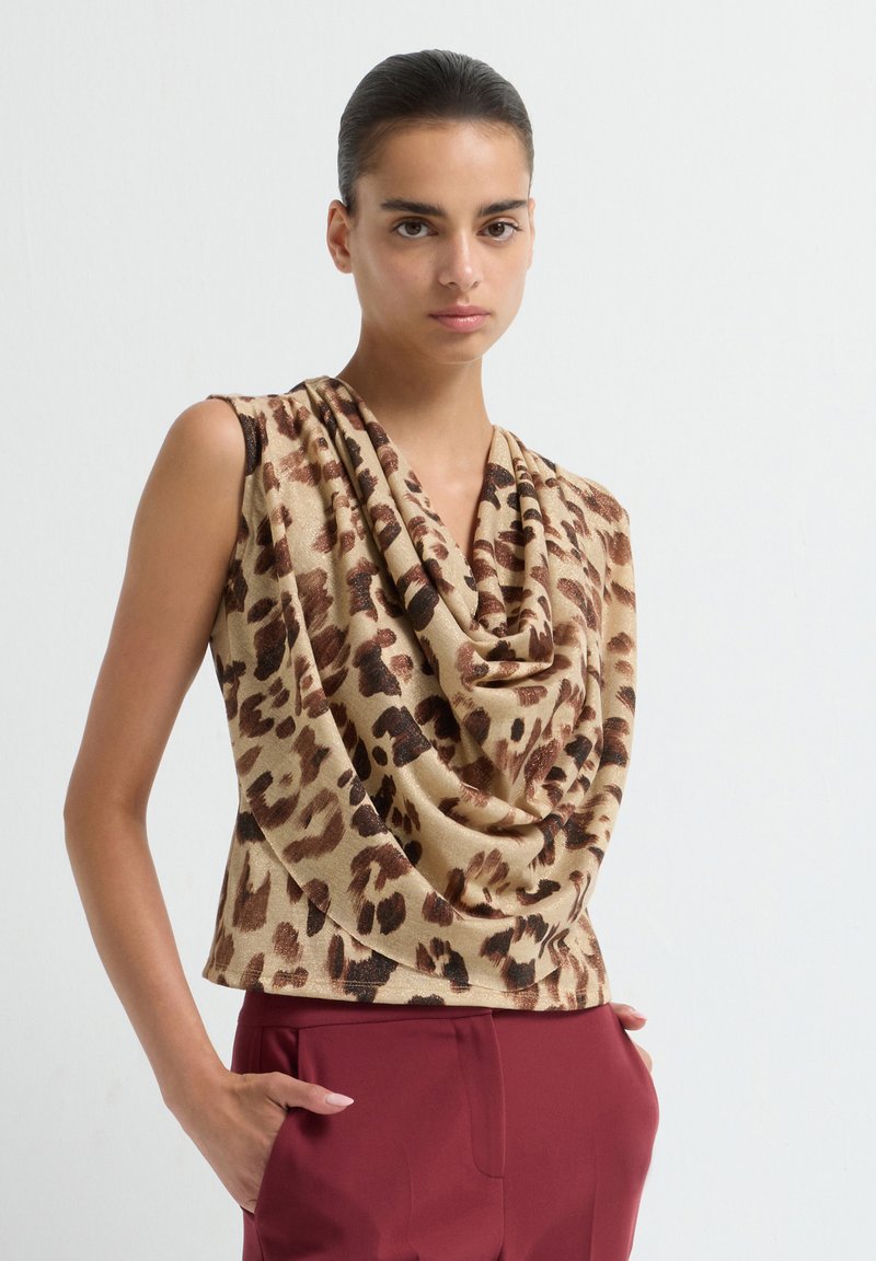 Vrouw in een mouwloze beige top met bruine luipaardprint en gedrapeerde halslijn, gecombineerd met hooggewaisted bordeauxrode broek, staand tegen een effen achtergrond.