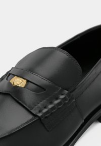 Črne usnjene loaferice z zlatim emblemom, ki prikazuje obraz z očali, podrobno šivanje ob traku in območju vamp.