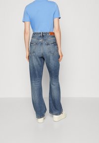 Person som bär högmidjade blå denimjeans med vida ben, en ljusblå kortärmad skjorta och vita plattformssneakers, stående vänd bort.