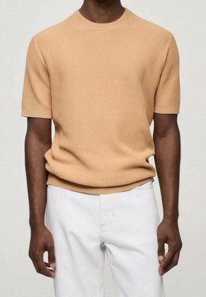 Homme portant un pull en maille texturée beige à manches courtes associé à un pantalon blanc, debout devant un fond clair uni.