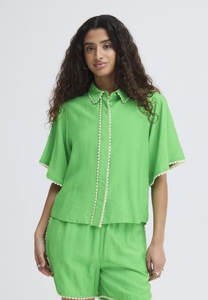 Frau mit langen lockigen Haaren, die ein hellgrünes Kurzarmshirt und passende Shorts mit weißem gewelltem Saum trägt, steht vor einem schlichten Hintergrund.