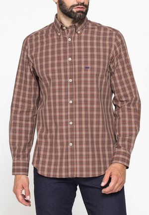 Uomo che indossa una camicia a quadri marrone, bianca e blu navy a maniche lunghe con colletto abbottonato e pantaloni blu scuro.