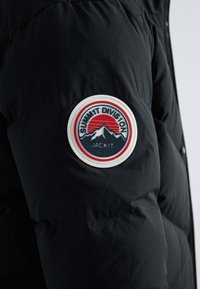 Doudoune noire avec un patch circulaire blanc sur la manche gauche, présentant l'inscription "SUMMIT DIVISION", des montagnes et un dessin de soleil rouge.