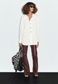 Camicia bianca con bottoni e maniche lunghe, pantaloni a campana in finta pelle marrone, borsa nera e maglione a righe. La modella indossa occhiali da sole.