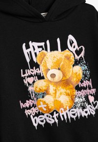 Sudadera negra con un gráfico de un oso de dibujos animados. El oso es esponjoso y marrón claro con una nariz marrón, rodeado de texto colorido y diseños salpicados.