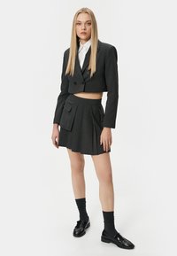 Grijze, cropped dubbele blazer met notch-revers, gecombineerd met een geplooide minirok met hoge taille en een zijzak. Zwarte loafers en donkere sokken.