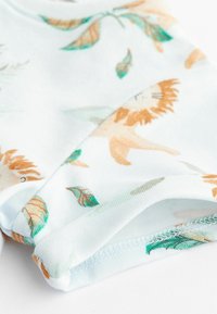 Tissu blanc avec une couture, présentant un motif de lions orange et de feuilles vertes, montré en gros plan.