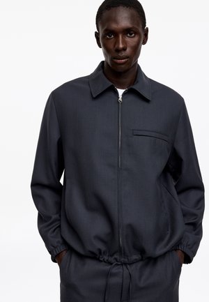 Homme à la peau foncée portant une veste zippée gris foncé avec des poignets élastiques et un ourlet à cordon, debout les mains dans les poches sur un fond blanc.