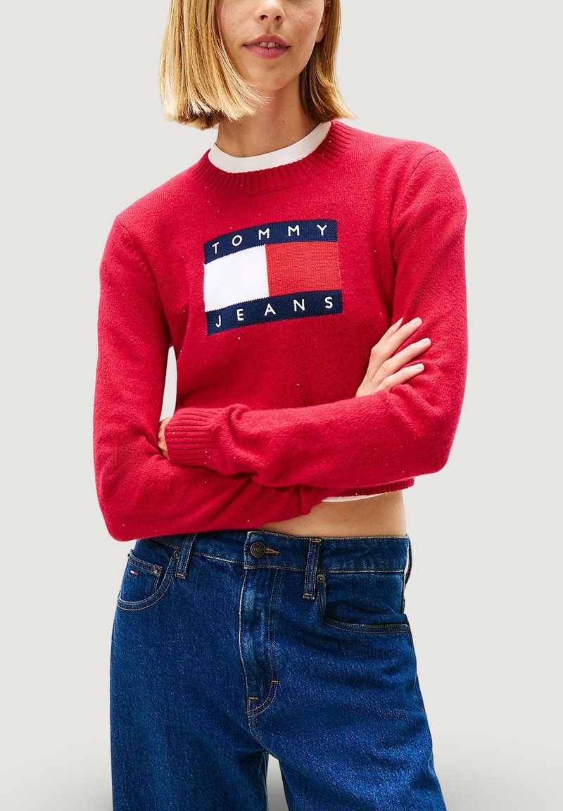 Roter Strickpullover mit Rundhalsausschnitt, mit einem rechteckigen Logo-Patch in Weiß, Rot und Marineblau. Kombiniert mit blauen Jeans.