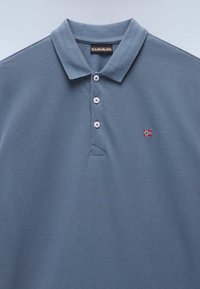 Lyseblå poloshirt med teksturert stoff, med en tre-knappers åpning og et lite brodert rødt og hvitt flagg på brystet.