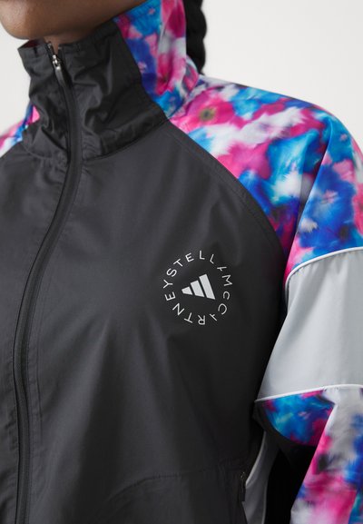 Veste coupe-vent noire avec des épaules à motifs floraux colorés, fermeture éclair à l'avant et accent de logo sur la poitrine. Texture lisse avec des détails contrastants.