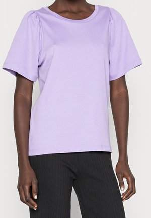 Personne portant un haut violet clair à manches courtes avec de légers fronces aux épaules et un pantalon noir côtelé sur un fond uni.