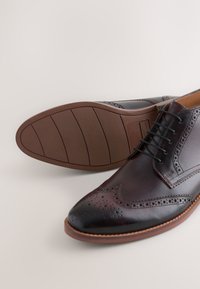 Braune Lederschuhe mit Brogue-Detailierung, spitzer Zehe und brauner Gummisohle. Die Schnürsenkel sind schwarz, mit glatter Textur und poliertem Finish.