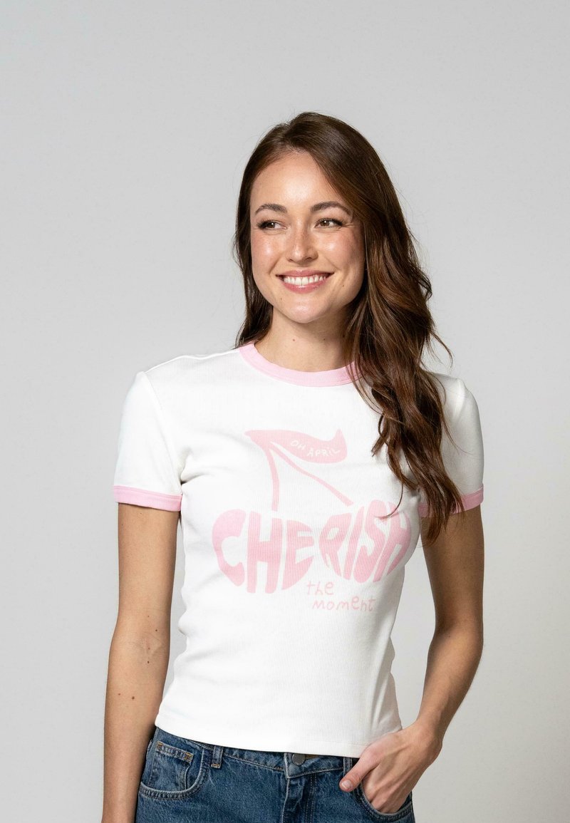 T-shirt blanc à manches courtes avec des accents rose clair, arborant un imprimé graphique avec le texte "CHÉRISSEZ le moment" et un design de chaussure à talon.