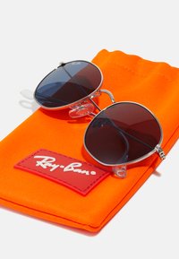 Occhiali da sole rotondi in metallo con lenti scure, poggiati su un sacchettino di stoffa arancione con un'etichetta del logo Ray-Ban rossa.