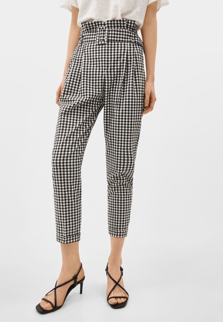 Femme portant un pantalon ajusté taille haute à carreaux vichy noir et blanc et des sandales à talons noires à fines lanières, debout devant un fond uni.