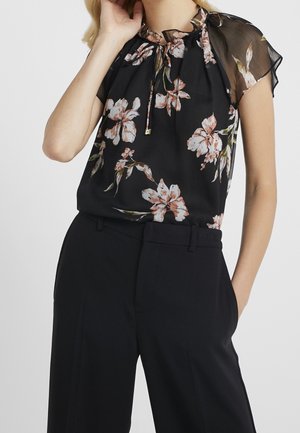 Kvinde iført en sort bluse med blomsterprint med korte gennemsigtige ærmer og højtsiddende sorte bukser, poserer med én hånd tæt på ansigtet.
