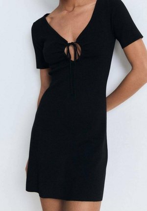 Kobieta ubrana w dopasowaną czarną sukienkę z krótkim rękawem, z wycięciem w kształcie klucza na przodzie i wiązaniem, stojąca na jednolitym jasnym tle.