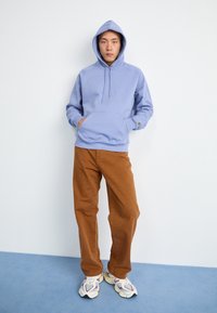 Haut à capuche en coton bleu clair avec une poche avant et un cordon de serrage, associé à un pantalon marron et des baskets multicolores chunky.