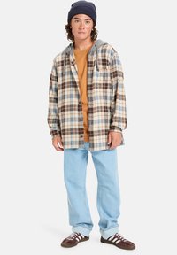 Camisa de cuadros en beige, marrón y azul; llevada sobre una camiseta naranja. Jeans de mezclilla azul claro y zapatillas marrones con cordones blancos.