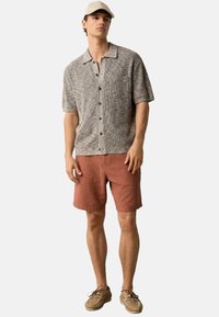 Kortærmet skjorte med knaplukning i struktureret beige med en brystlomme, parret med rustfarvede shorts og tan sneakers. Kasketter båret let på skrå.
