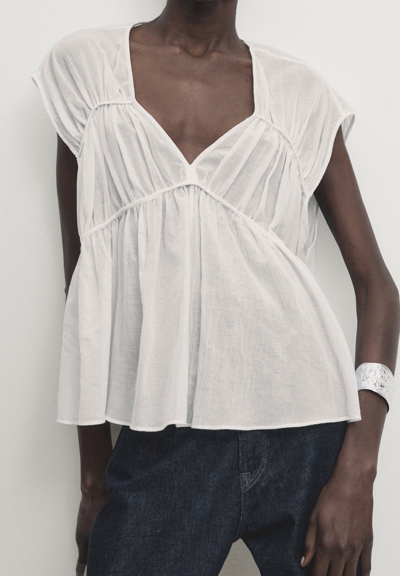 Blouse blanche, ample, avec un col en V et des plis froncés. Manches courtes et légère évasement à l'ourlet. Portée avec un jean en denim foncé.