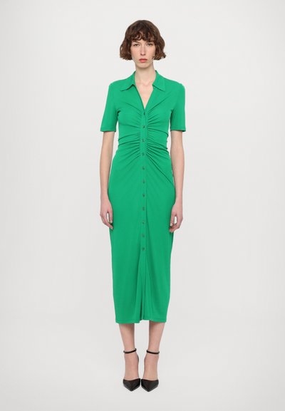 Vestido midi verde en tela elástica, con botones en la parte delantera, mangas cortas y detalles fruncidos en la cintura. Combinado con tacones negros puntiagudos.