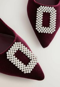 Par de zapatos de terciopelo burdeos con punta puntiaguda, adornados con hebillas rectangulares de piedras brillantes en la parte superior.