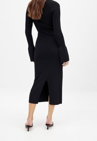 Robe noire côtelée avec manches longues et poignets évasés. Longueur midi et fente au dos pour faciliter les mouvements. Texture lisse.