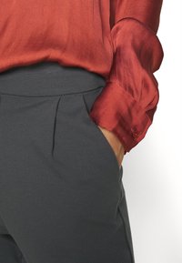 Sidenblus i röd färg med subtil lyster och knäppning vid ärmarna, kombinerad med gråbyxor som har en mjuk textur och veck fram.