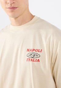 Beige bomull T-skjorte med en rød grafikk som leser "NAPOLI ITALIA" og en pizza-illustrasjon på venstre brystområde. Myk, uformell passform.