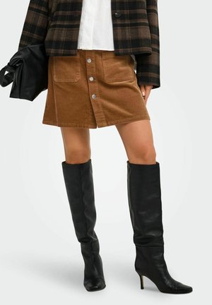 Femme portant une jupe marron en velours côtelé à boutons sur le devant, des bottes noires à talons hauts jusqu'aux genoux, une veste à carreaux, tenant un sac à main noir.