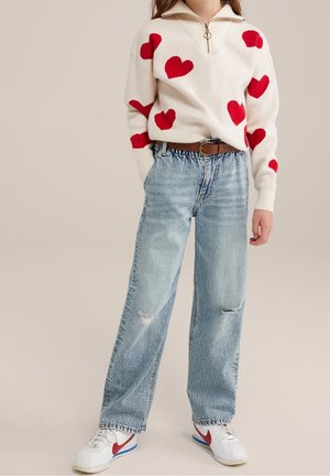 Kvinde iført hvid sweater med røde hjerter, lyseblå revnede jeans, brunt bælte og hvide sneakers med rødt swoosh-logo.