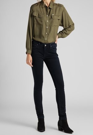 Osoba w oliwkowej koszuli zapinanej na guziki, ciemnych obcisłych dżinsach i czarnych botkach, stojąca na tle jednolitego szarego tła.