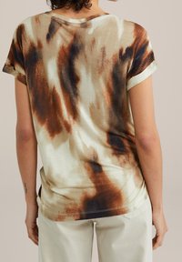 Krótki rękaw bluzki w kolorze kremowym z brązowym i czarnym abstrakcyjnym wzorem, z podwiniętymi mankietami i luźnym krojem, wykonana z miękkiego materiału.