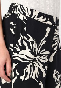 Pantaloni din material negru, cu un model floral mare alb, având o talie plisată și buzunare laterale. Textură moale, croială lejeră.