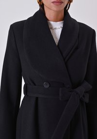 Schwarzer Wollmantel mit großem Kragen, doppelten Knöpfen und einem gebundenen Gürtel in der Taille. Texturiertes Gewebe mit strukturiertem Design.