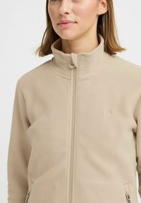 Beige fleece-takki, jossa on korkea kaulus, täysivetoketju ja sivutaskut. Ominaisuuksiin kuuluu teksturoitu pinta ja pieni logo rinnassa.