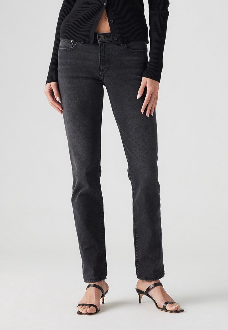 Femme portant un jean noir ajusté, un haut noir boutonné et des sandales à talons noires à brides, debout devant un fond clair uni.